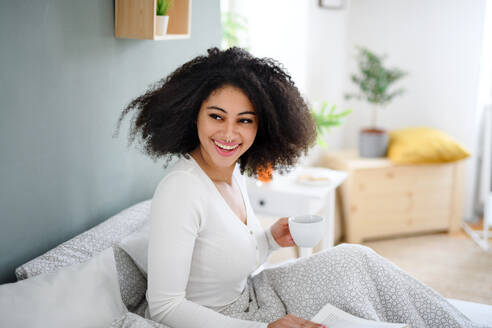 Close-up portrait of happy young woman with book and coffee indoors, relaxing in bed. - HPIF08727