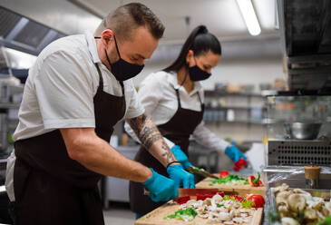 A chef and cook with face masks cutting vegetables indoors in restaurant kitchen, coronavirus concept. - HPIF08775