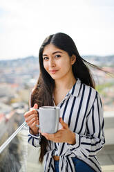 Ein Porträt einer jungen Frau mit Kaffee auf einem Balkon in der Stadt, die in die Kamera schaut. - HPIF08777