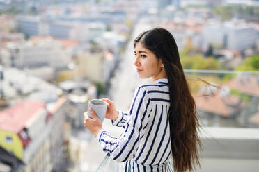 Ein Porträt einer jungen Frau mit Kaffee, die auf einem Balkon in der Stadt steht und auf die Stadtlandschaft blickt. - HPIF08778