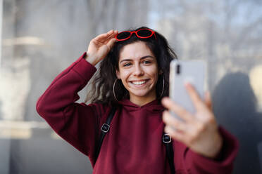 A young woman captures a selfie while standing on a city street, using her smartphone to snap the perfect shot - HPIF08805