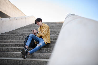 A portrait of young man sitting on staircase outdoors in city, using smartphone - HPIF08817