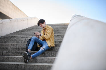 Porträt eines jungen Mannes, der auf einer Treppe in der Stadt sitzt und ein Smartphone benutzt - HPIF08817