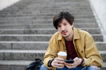 A portrait of young man sitting on staircase outdoors in city, using smartphone - HPIF08818