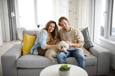 A happy young couple in love with dog sitting on sofa indoors at home, looking at camera. - HPIF08821