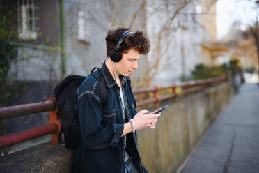 A portrait of young man commuter with headphones standing outdoors in city, using smartphone. - HPIF08854