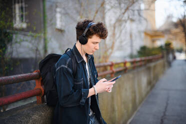 Ein Porträt eines jungen Mannes, der mit Kopfhörern im Freien in der Stadt steht und sein Smartphone benutzt. - HPIF08854