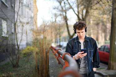 A portrait of young man commuter with headphones walking outdoors in city, using smartphone. - HPIF08855