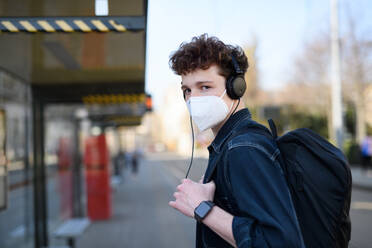 A portrait of young man commuter standing on bus stop outdoors in city, coronavirus concept. - HPIF08859