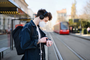 A portrait of young man commuter standing on bus stop outdoors in city, coronavirus concept. - HPIF08860