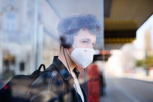 A portrait of young man commuter standing on bus stop outdoors in city, coronavirus concept. - HPIF08861
