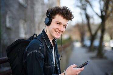 Ein Porträt eines jungen Mannes, der mit Kopfhörern im Freien in der Stadt steht und sein Smartphone benutzt. - HPIF08865