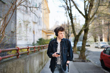 A portrait of young man commuter with headphones walking outdoors in city, using smartphone. - HPIF08866