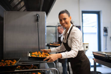 A happy female cook looking at camera and holding tray with baked pumpkin pieces in commercial kitchen. - HPIF08873