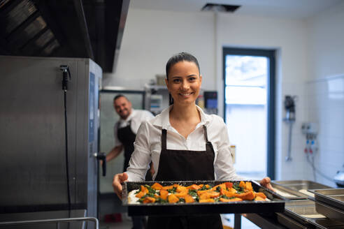 A happy female cook looking at camera and holding tray with baked pumpkin pieces in commercial kitchen. - HPIF08875