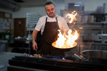 Ein professioneller Koch bereitet eine Mahlzeit zu, flambiert drinnen in einer Restaurantküche. - HPIF08927