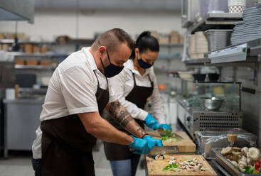 A chef and cook with face masks cutting vegetables indoors in restaurant kitchen, coronavirus concept. - HPIF08935