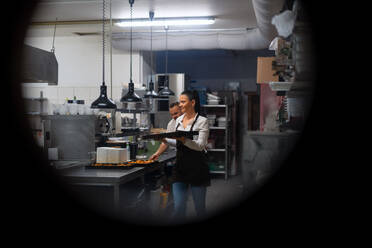 Ein glücklicher Koch und eine Köchin bei der Arbeit an ihren Gerichten in der Restaurantküche, aufgenommen durch das Fenster einer Rundtür. - HPIF08941