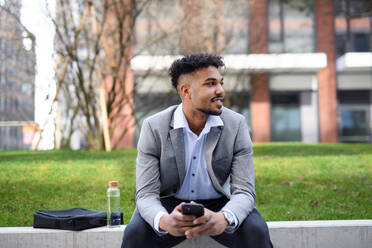 A portrait of young man student sitting outdoors in city, using smartphone. - HPIF08958