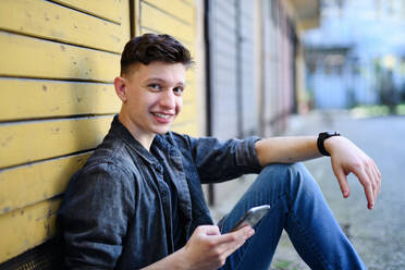 Ein Porträt eines jungen Mannes mit Smartphone, der draußen auf einer Straße in der Stadt sitzt und in die Kamera schaut. - HPIF08971