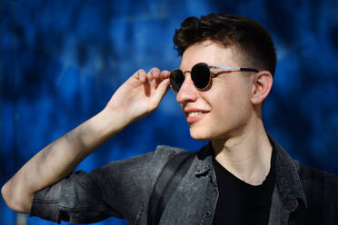 A portrait of young man with sunglasses standing outdoors against blue background. - HPIF08972