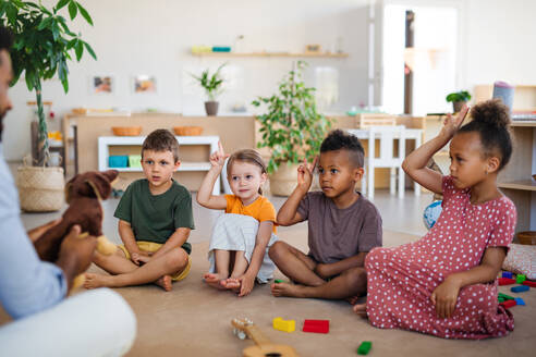 Eine Gruppe von kleinen Kindergartenkindern mit einem männlichen Lehrer, der im Klassenzimmer auf dem Boden sitzt und die Hände hebt. - HPIF08997
