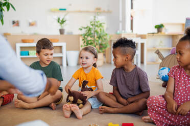 Eine Gruppe von kleinen Kindergartenkindern, die in einem Klassenzimmer auf dem Boden sitzen und dem Lehrer zuhören. - HPIF08999