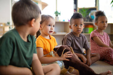 A group of small nursery school children sitting on floor indoors in classroom, listening to teacher. - HPIF09005