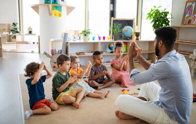 Eine Gruppe von kleinen Kindergartenkindern mit einem männlichen Lehrer, die in einem Klassenzimmer auf dem Boden sitzen und spielen. - HPIF09006