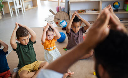 Eine Gruppe von kleinen Kindergartenkindern mit einem männlichen Lehrer, die in einem Klassenzimmer auf dem Boden sitzen und spielen. - HPIF09007