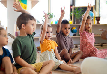 Eine Gruppe von kleinen Kindergartenkindern, die in einem Klassenzimmer auf dem Boden sitzen und die Hände heben. - HPIF09012