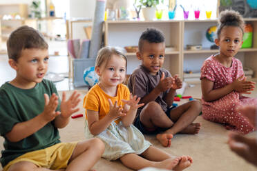 Eine Gruppe kleiner Kindergartenkinder sitzt in einem Klassenzimmer auf dem Boden und klatscht. - HPIF09014