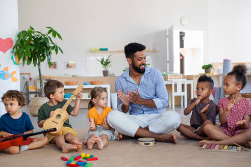 Eine Gruppe von kleinen Kindergartenkindern mit einem männlichen Lehrer, die in einem Klassenzimmer auf dem Boden sitzen und Musikinstrumente spielen. - HPIF09018