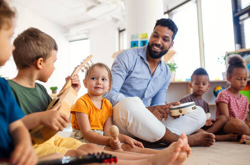 Eine Gruppe von kleinen Kindergartenkindern mit einem männlichen Lehrer sitzt auf dem Boden in einem Klassenzimmer und spielt Musikinstrumente. - HPIF09019