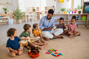 Eine Gruppe von kleinen Kindergartenkindern mit einem männlichen Lehrer, die in einem Klassenzimmer auf dem Boden sitzen und Musikinstrumente spielen. - HPIF09021
