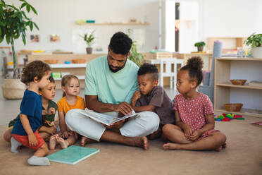 Eine Gruppe von kleinen Kindergartenkindern mit einem männlichen Lehrer, der in einem Klassenzimmer auf dem Boden sitzt und ein Märchenbuch liest. - HPIF09022