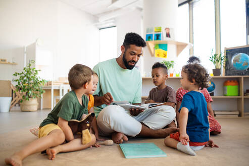 Eine Gruppe von kleinen Kindergartenkindern mit einem männlichen Lehrer, der in einem Klassenzimmer auf dem Boden sitzt und Unterricht hat. - HPIF09023