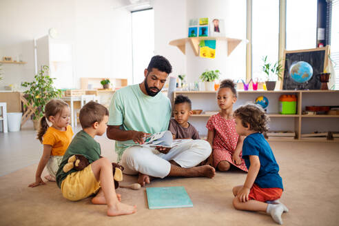 Eine Gruppe von kleinen Kindergartenkindern mit einem männlichen Lehrer, der in einem Klassenzimmer auf dem Boden sitzt und Unterricht hat. - HPIF09025