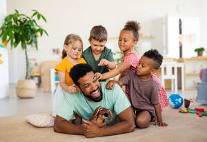 Eine Gruppe von kleinen Kindergartenkindern mit einem männlichen Lehrer, die in einem Klassenzimmer auf dem Boden sitzen und spielen. - HPIF09027