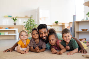 Eine Gruppe von kleinen Kindergartenkindern mit einem männlichen Lehrer auf dem Boden in einem Klassenzimmer, der in die Kamera schaut. - HPIF09029