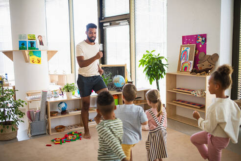 Eine Gruppe von kleinen Kindergartenkindern mit einem männlichen Lehrer, die in einem Klassenzimmer stehen und Übungen machen. - HPIF09034