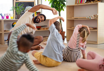 Eine Gruppe von kleinen Kindergartenkindern mit einem männlichen Lehrer sitzt auf dem Boden in einem Klassenzimmer und macht Übungen. - HPIF09035