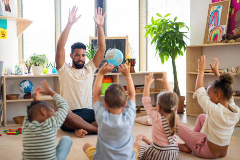 Eine Gruppe von kleinen Kindergartenkindern mit einem männlichen Lehrer sitzt auf dem Boden in einem Klassenzimmer und macht Übungen. - HPIF09036