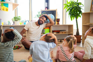 Eine Gruppe von kleinen Kindergartenkindern mit einem männlichen Lehrer sitzt auf dem Boden in einem Klassenzimmer und macht Übungen. - HPIF09037