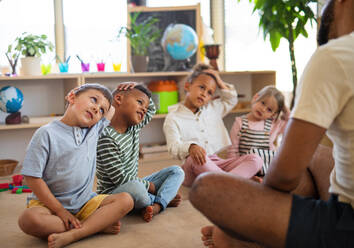 Eine Gruppe von kleinen Kindergartenkindern mit einem männlichen Lehrer sitzt auf dem Boden in einem Klassenzimmer und macht Übungen. - HPIF09038