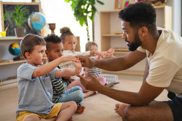 Eine Gruppe von kleinen Kindergartenkindern mit einem männlichen Lehrer sitzt auf dem Boden in einem Klassenzimmer und macht Übungen. - HPIF09039