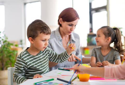 Eine Gruppe kleiner Kindergartenkinder mit einer Lehrerin in einem Klassenzimmer beim Malen. - HPIF09059