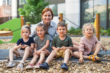 Eine Gruppe von kleinen Kindergartenkindern mit einer Lehrerin, die auf einem Spielplatz im Freien sitzen und in die Kamera schauen. - HPIF09069