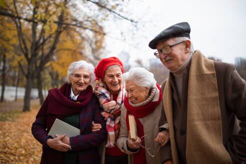 Eine Gruppe glücklicher älterer Freunde mit Büchern auf einem Spaziergang im Park im Herbst, die sich unterhalten und lachen - HPIF09095