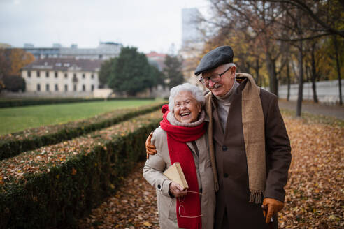 A happy senior couple on walk outdoors in town park in autumn, embracing and laughing. - HPIF09101